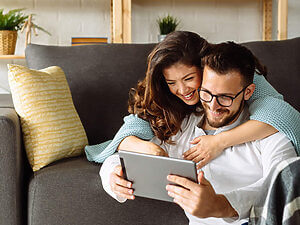 Ein Paar lächelt, während es gemeinsam auf einem Tablet einen 360-Grad-Immobilienrundgang anschaut und dabei auf dem Sofa im Wohnzimmer sitzt.
