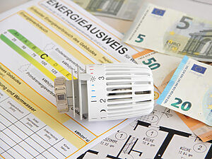 Ein Thermostat auf einem Hausgrundriss neben Euro-Scheinen und einem Energieausweis, auf Wunsch von uns als Ihrem Immobilienmakler.