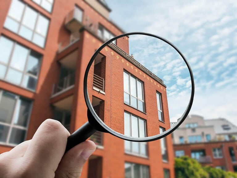 Eine in der Hand gehaltene Lupe vergrößert einen Teil eines modernen roten Mehrfamilienhauses in Hiddenhausen vor einem blauen Himmel mit Wolken.