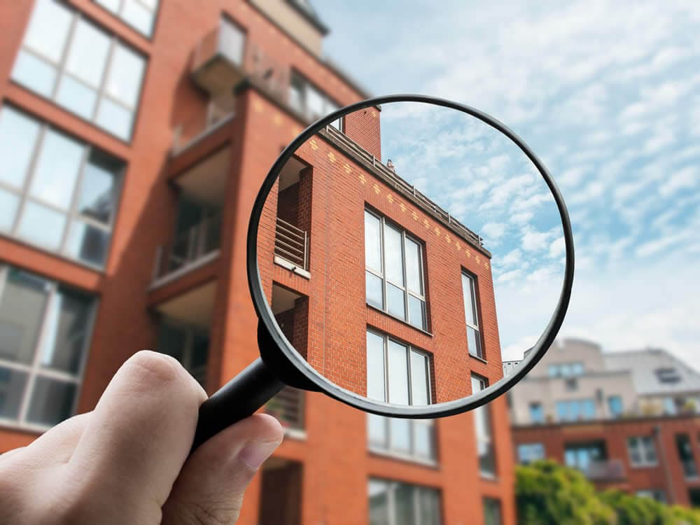Eine in der Hand gehaltene Lupe vergrößert einen Teil eines modernen roten Mehrfamilienhauses in Hiddenhausen vor einem blauen Himmel mit Wolken. Eine in der Hand gehaltene Lupe vergrößert einen Teil eines modernen roten Mehrfamilienhauses in Hiddenhausen vor einem blauen Himmel mit Wolken.