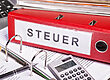Roter Ordner mit der Aufschrift „Steuer“ auf einem Schreibtisch, umgeben von Papierkram und einem Taschenrechner, als Symbol für die Steuern, die in Zusammenhang mit einer Immobilie anfallen können.