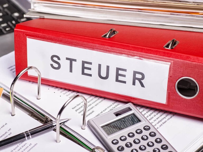 Roter Ordner mit der Aufschrift „Steuer“ auf einem Schreibtisch, umgeben von Papierkram und einem Taschenrechner, als Symbol für die Steuern, die in Zusammenhang mit einer Immobilie anfallen können.