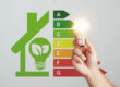 Eine Hand hält eine leuchtende Glühbirne vor einer Energieeffizienz-Tabelle und einem grünen Haus-Symbol.