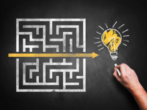 Eine Hand zeichnet einen gelben Pfeil, der ein Kreidelabyrinth auf einer Tafel durchschneidet und an einer Glühbirne endet - ein Symbol dafür, wie man den für sich passenden und qualifizierten Immobilienmakler findet und auswählt.