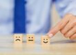 Eine Hand zeigt auf einen Holzklotz mit einem lächelnden Gesicht, der neben Klötzen mit neutralen und traurigen Gesichtern auf einem Tisch liegt - ein Symbol beim Immobilienverkauf: warum der erste Eindruck so wichtig ist.