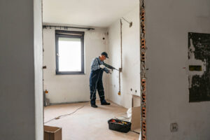 Eine Person in einem Blaumann bohrt mit einer Bohrmaschine in die Wand eines teilweise renovierten Raums mit verstreuten Werkzeugen und Materialien, um den Raum für die Sanierung und einen anschließenden Immobilienverkauf vorzubereiten.