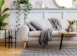 Home Staging für den Hausverkauf: Ein modernes Wohnzimmer wird durch ein hellbeiges Sofa, eine graue Decke, einen Couchtisch aus Holz, Pflanzen, gerahmte Kunst und einen runden Spiegel an einer getäfelten Wand deutlich emotionaler dargestellt als ein leerer Raum.