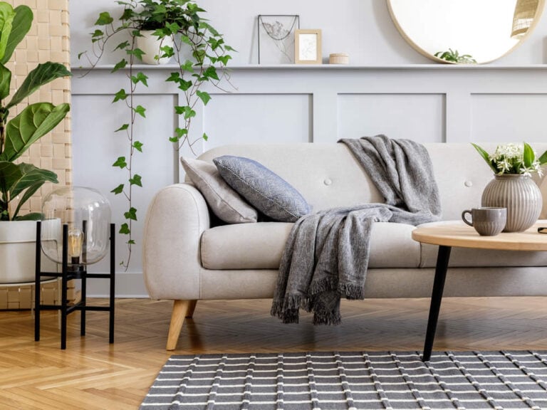 Home Staging für den Hausverkauf: Ein modernes Wohnzimmer wird durch ein hellbeiges Sofa, eine graue Decke, einen Couchtisch aus Holz, Pflanzen, gerahmte Kunst und einen runden Spiegel an einer getäfelten Wand deutlich emotionaler dargestellt als ein leerer Raum. Home Staging für den Hausverkauf: Ein modernes Wohnzimmer wird durch ein hellbeiges Sofa, eine graue Decke, einen Couchtisch aus Holz, Pflanzen, gerahmte Kunst und einen runden Spiegel an einer getäfelten Wand deutlich emotionaler dargestellt als ein leerer Raum.