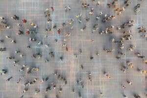 Luftaufnahme von verstreut positionierten Menschen, die über eine große offene Fläche mit schwachen, sich kreuzenden Linien gehen, die an die definierten Grenzen des Einzugsgebiets eines Immobilienmaklers erinnern.
