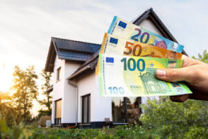 Eine Hand hält Euro-Banknoten vor einem modernen Haus in die Kamera – als Symbol für Immobilienfinanzierung und -förderung –, im Hintergrund sind Sonnenlicht und Grünflächen zu sehen.