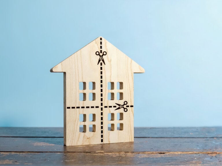 Ein ausgeschnittenes Holzhaus mit gepunkteten Linien und Scheren-Symbolen, die im Zusammenhang mit einer Teilungserklärung für eine Immobilie die Aufteilung symbolisieren, steht auf einer Holzfläche vor einem hellblauen Hintergrund.