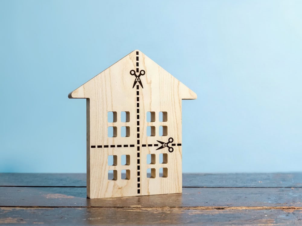 Ein ausgeschnittenes Holzhaus mit gepunkteten Linien und Scheren-Symbolen, die im Zusammenhang mit einer Teilungserklärung für eine Immobilie die Aufteilung symbolisieren, steht auf einer Holzfläche vor einem hellblauen Hintergrund. Ein ausgeschnittenes Holzhaus mit gepunkteten Linien und Scheren-Symbolen, die im Zusammenhang mit einer Teilungserklärung für eine Immobilie die Aufteilung symbolisieren, steht auf einer Holzfläche vor einem hellblauen Hintergrund.